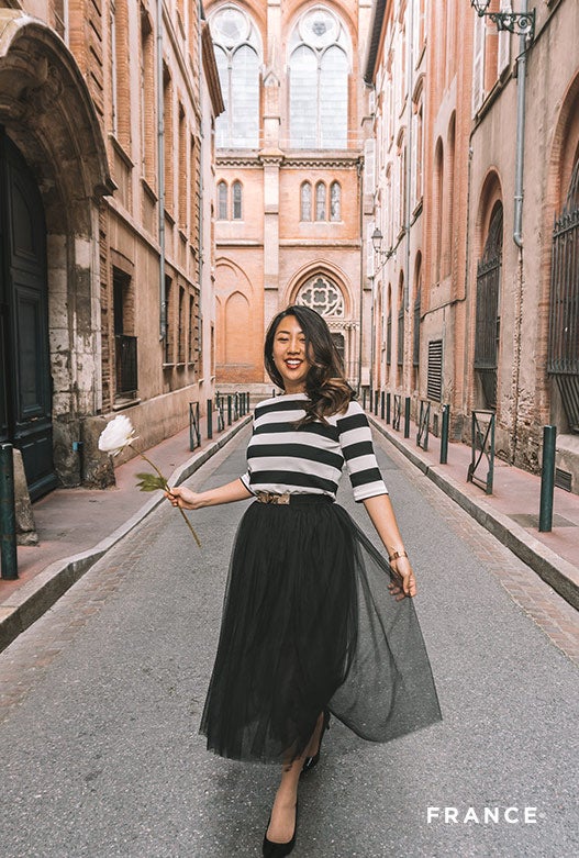 A woman in a black and white striped shirt and long black skirt holds a flower while walking down a bright alley.