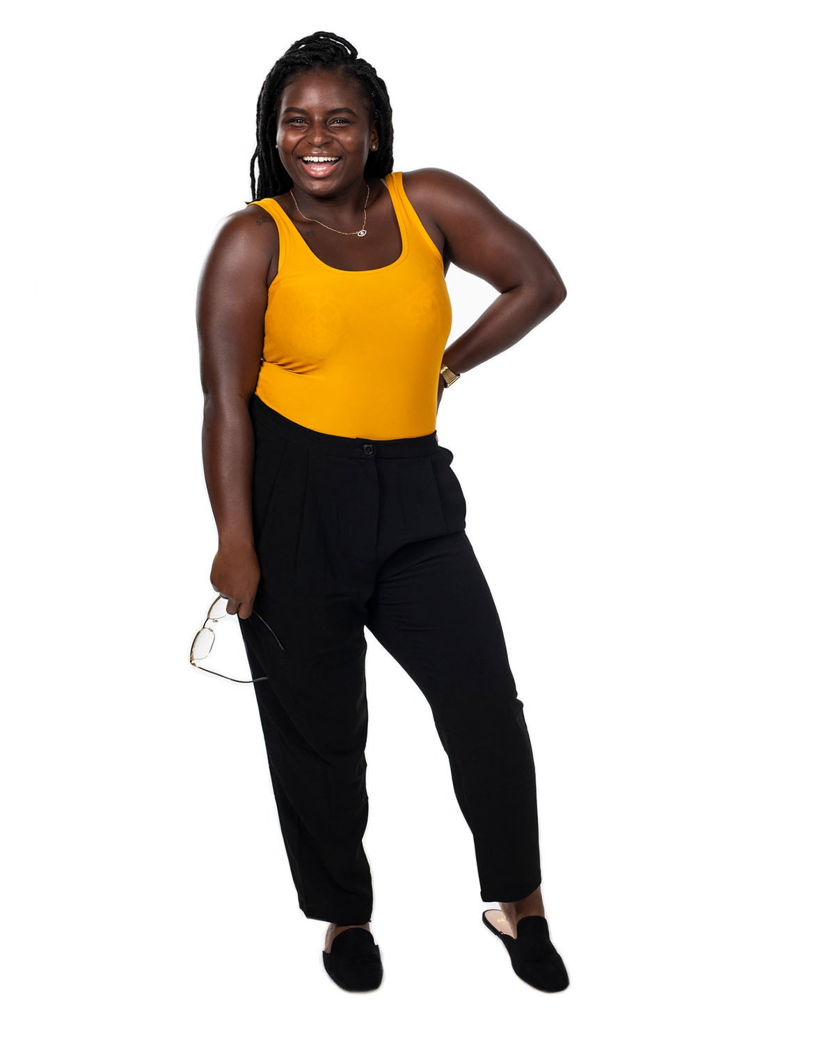 A woman in an orange tank top and black pants holds her glasses with one hand on her hip as she smiles.