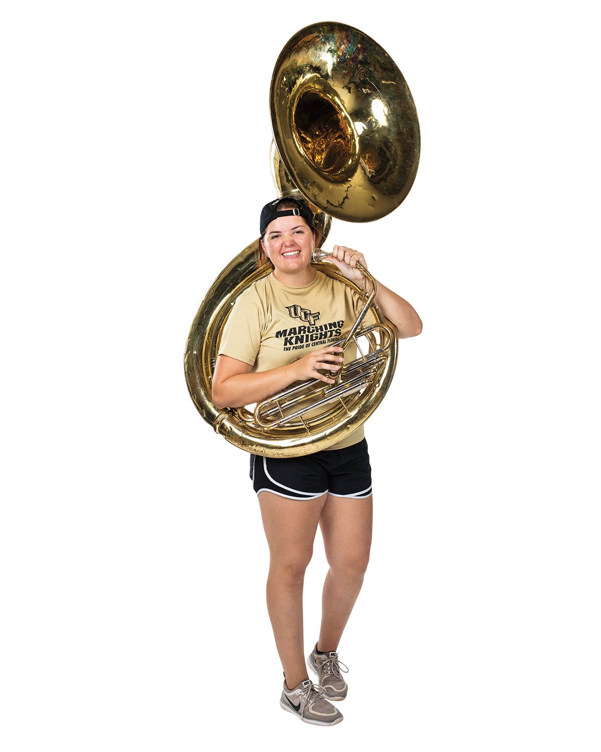 A woman in a yellow shirt and black shorts holds a tuba across her body as she smiles.