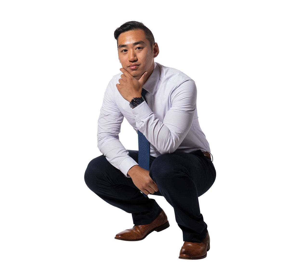 A man in a button down shirt, tie and slacks crouches down with his hand holding his chin.