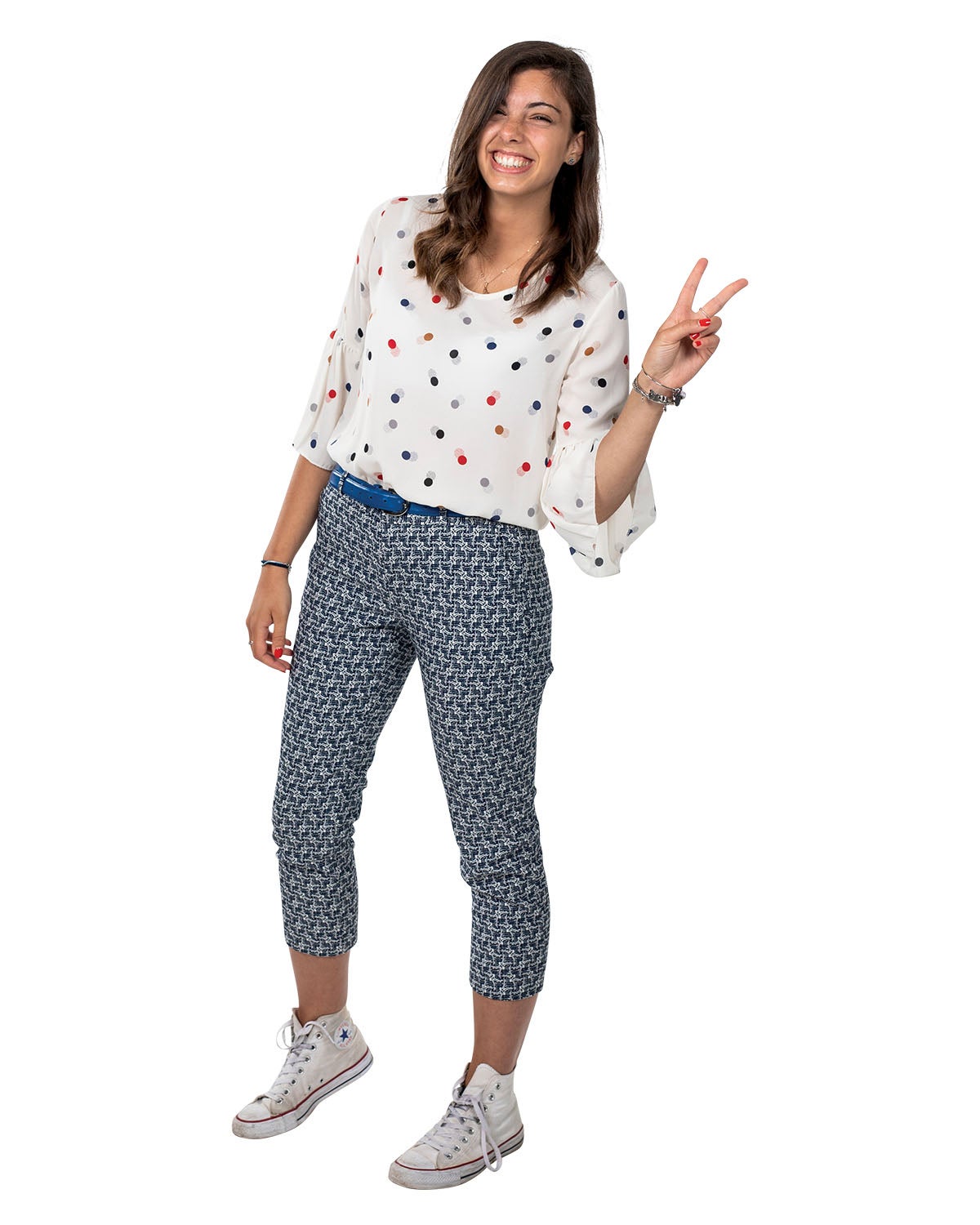 A woman in a white polka dot shirt and patterned pants holds up a peace sign with her fingers as she smiles.