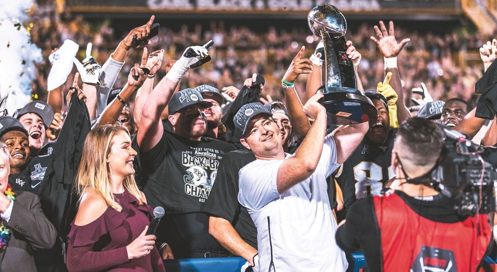 Josh Heupel, UCF football head coach holds up aac trophy with his team celebrating behind him