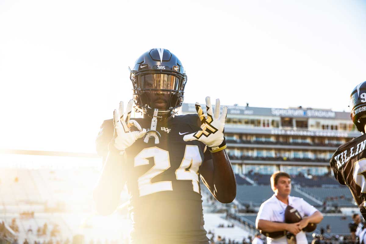 A football player wearing a black #24 jersey pops up his hands, wearing white gloves, with the sun setting in the background.