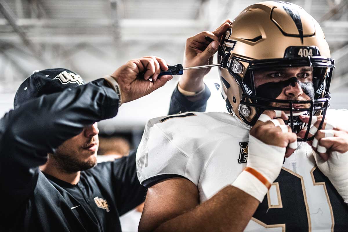 Player wearing a white jersey and gold football helmet helps get his helmet screwed on by a man in a black long sleeve shirt and black hat.