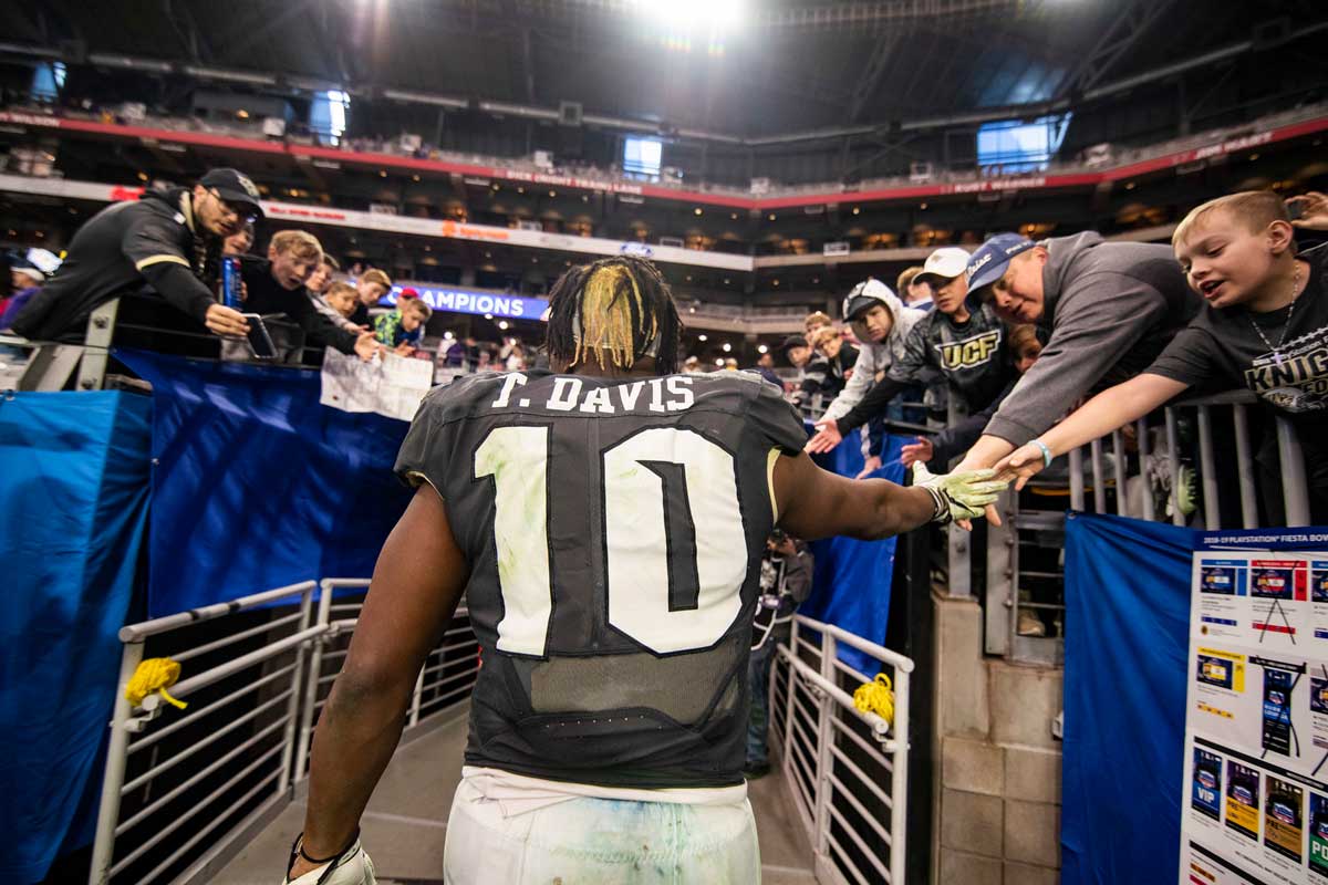 Football player wearing a black #10 jersey and white pants shakes hands with fans as he heads into tunnel under stadium.