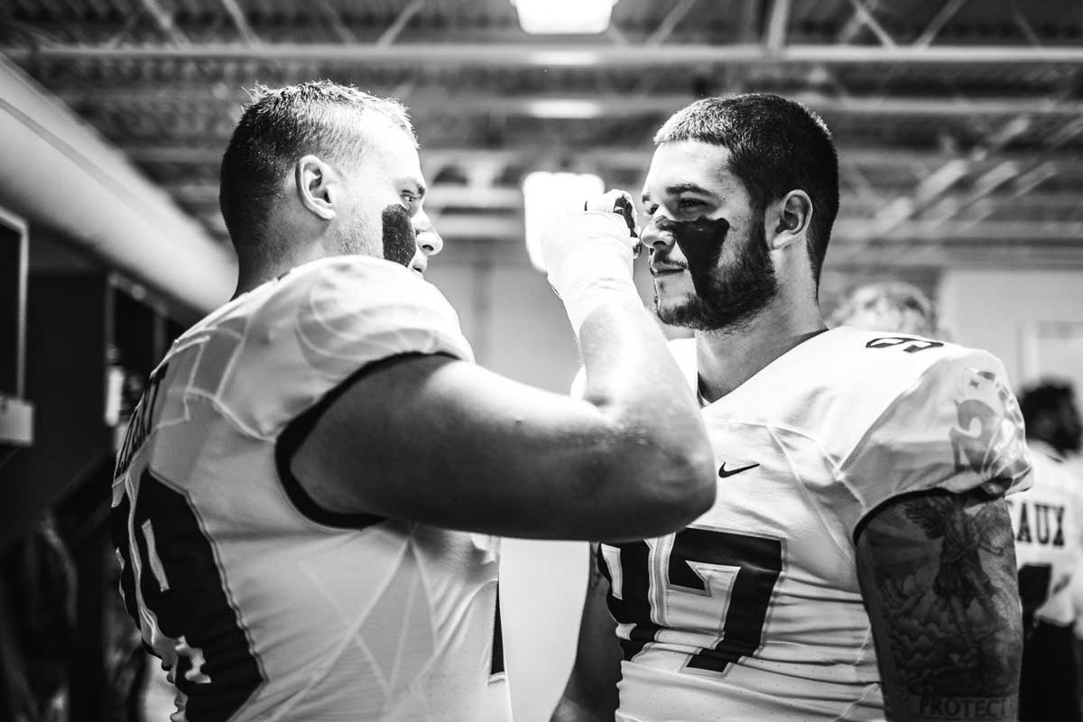 Black and white photo of one football player applying eye black to the face of another player in uniform