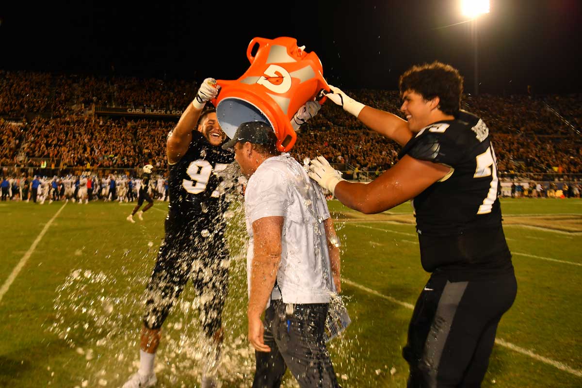 Two football players dump an orange Gatorade bucket of water on UCF head coach Josh Heupel on the field