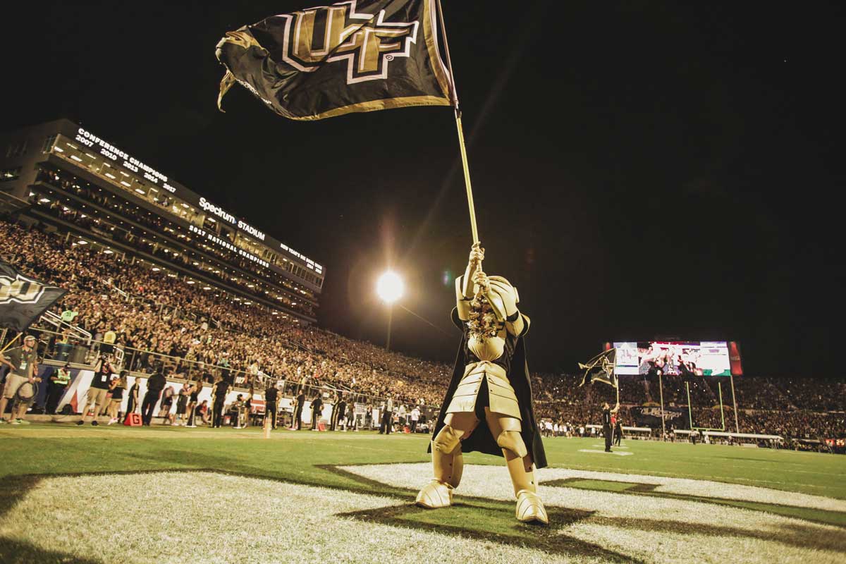 UCF mascot Knightro waves black and gold UCF flag on the field in front of Spectrum Stadium crowd at night