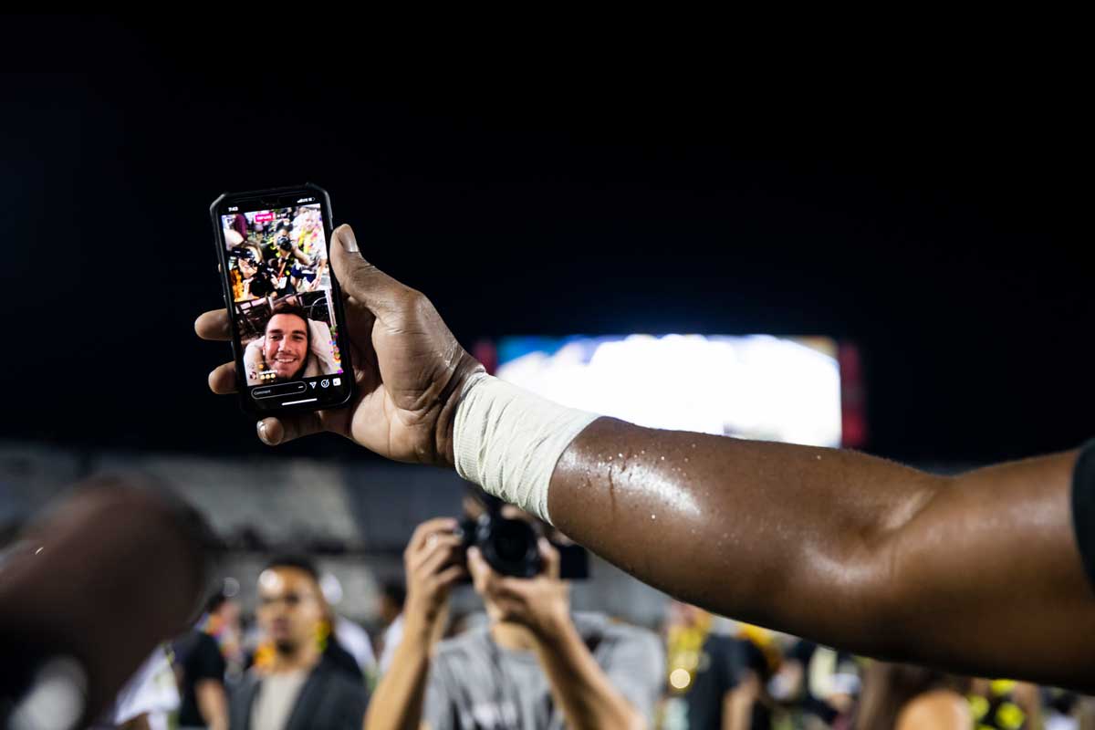 A hand on an outstretched arm grasps a cell phone FaceTiming in quarterback McKenzie Milton to the football field at night