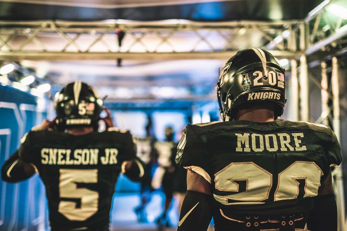 Photo shot from behind as two football players wearing black #5 and #20 jerseys walk in a tunnel under the stadium and head towards the light outside.