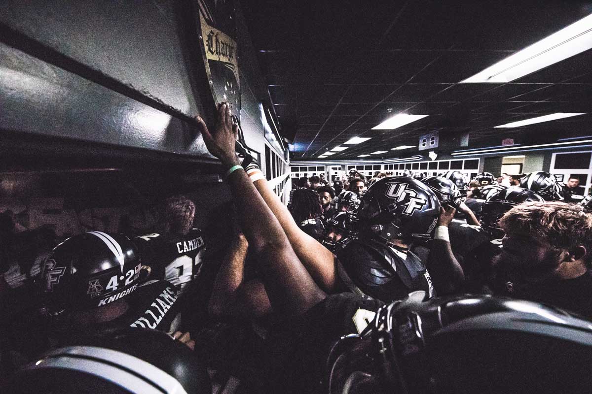 UCF players in full uniform reach up to touch a black and gold shield hanging in their locker room.