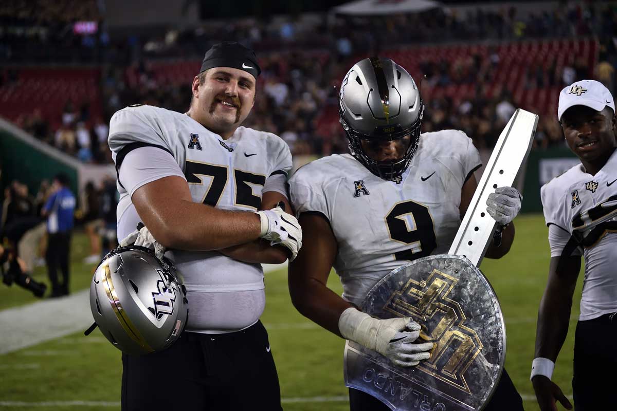 Two UCF football players pose with a silver trophy shaped like an interstate sign on the field