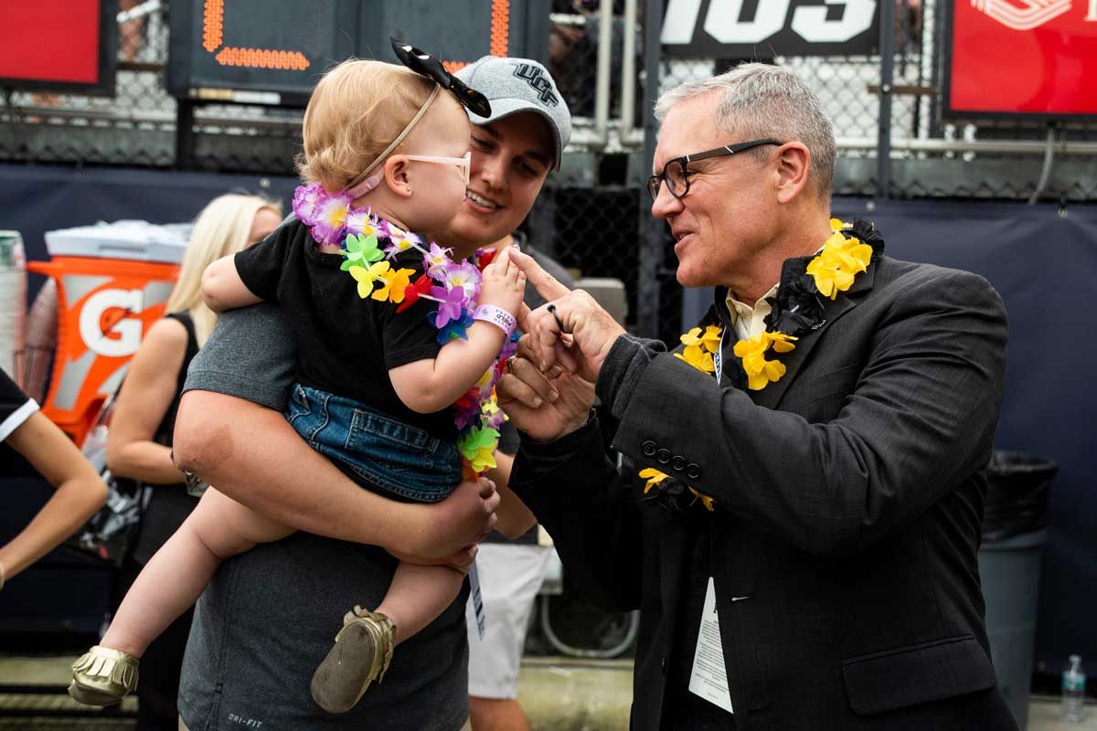 A man with short gray hair wearing black glasses and a black jacket and black and white lei shakes a toddler girl's hand as she is held by her father.