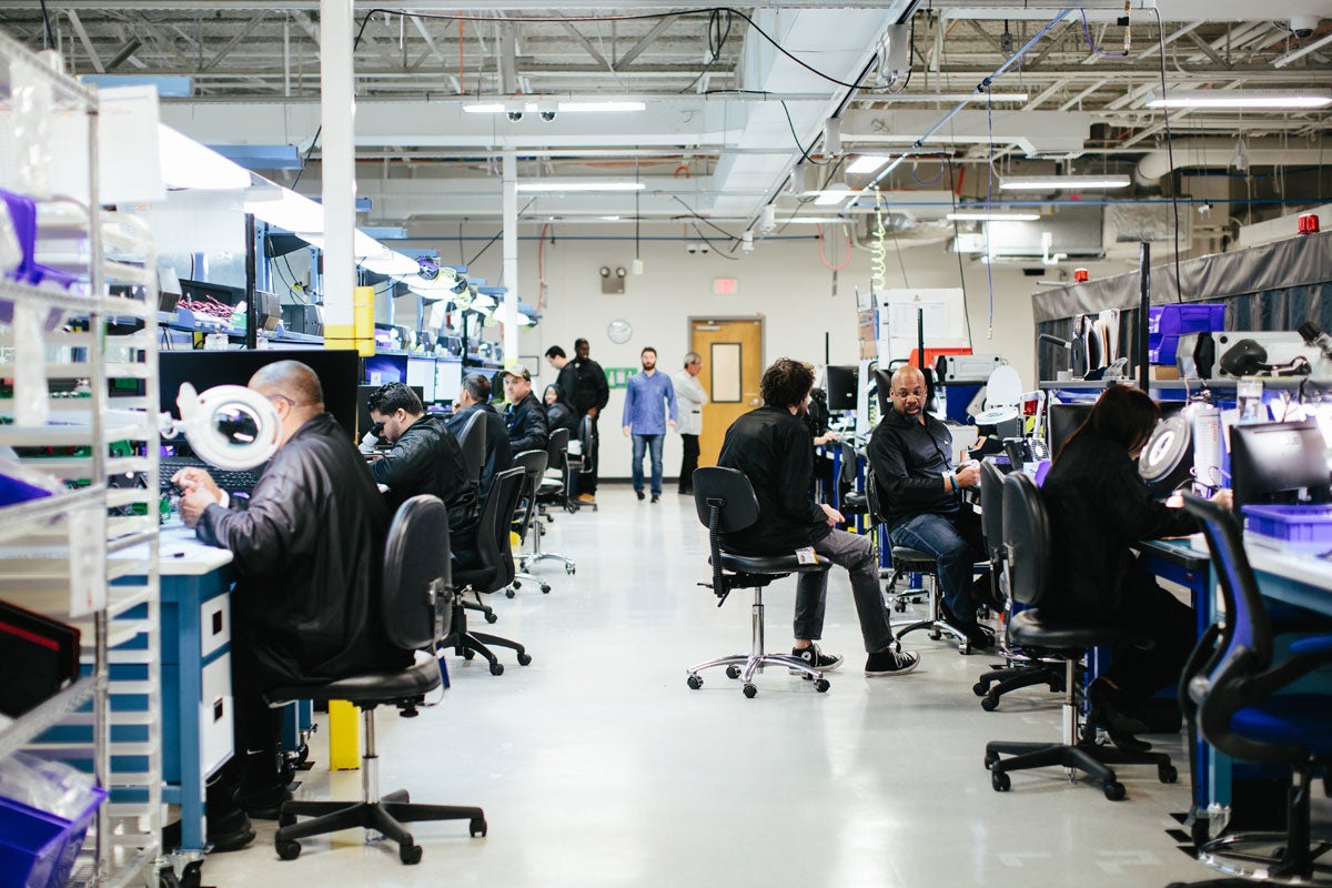 Two rows of workers sit in chairs while they tinker with electronics.