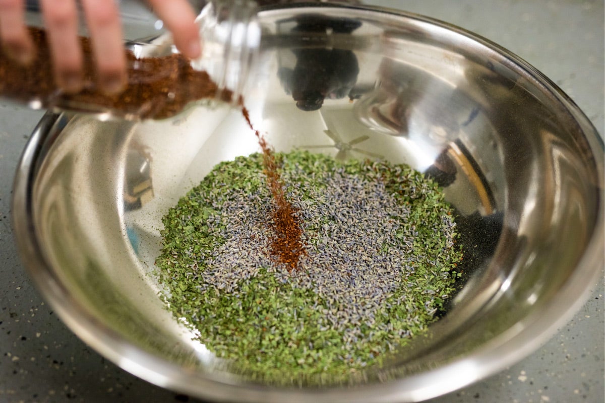 Someone pours a bottle of dried brown leaves into a metal bowl with green and purple dried leaves.