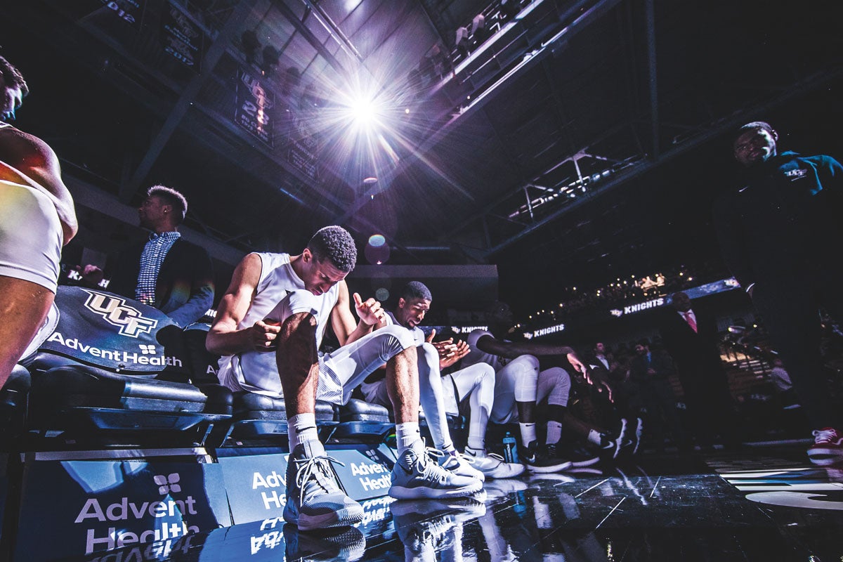 A group of basketball players sit on the sidelines before the start of a game.