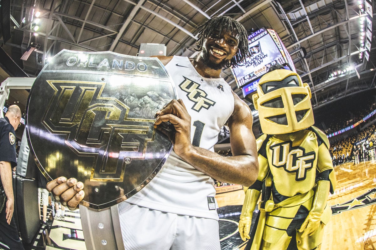 A UCF basketball player holds up a War-On-I-4 trophy with Knightro behind him.