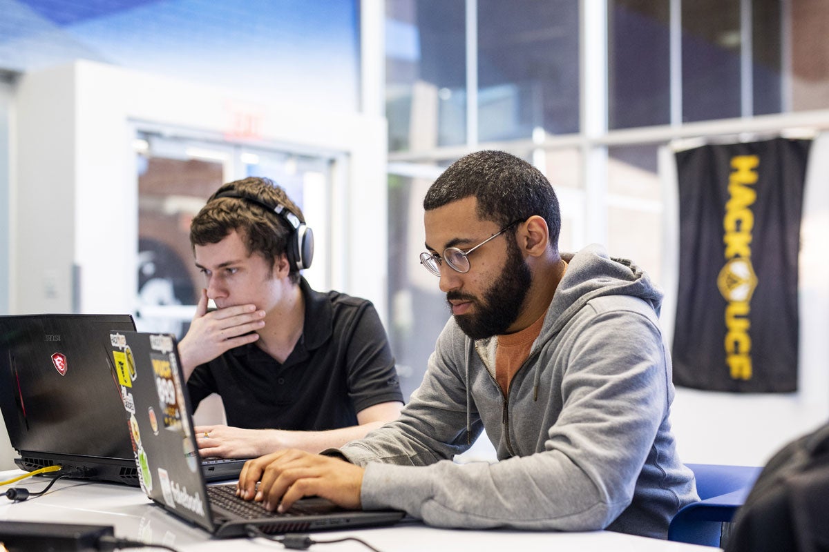 Two students work on their laptops while sitting at a whiteboard table.