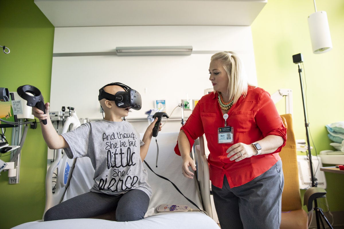 A young child in a hospital bed wears a VR headset while a woman stands nearby.