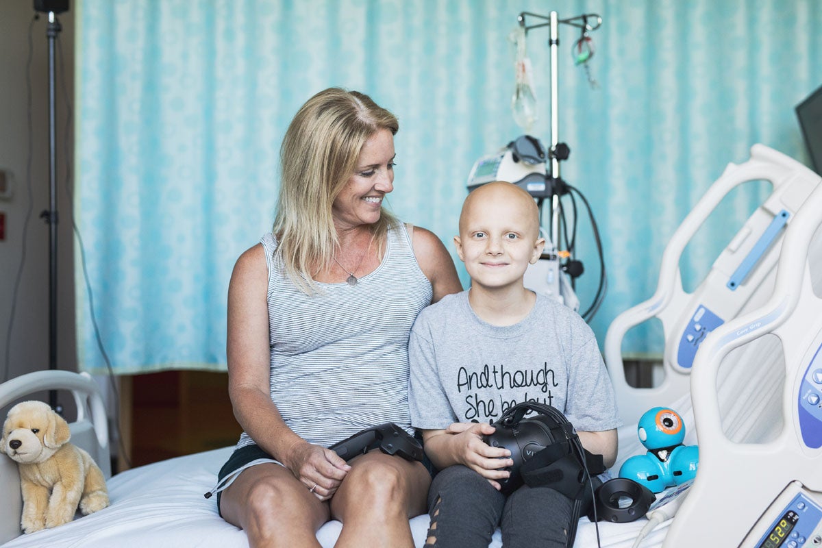 A young child in a hospital bed smiles and holds a VR headset while her mother looks at her.