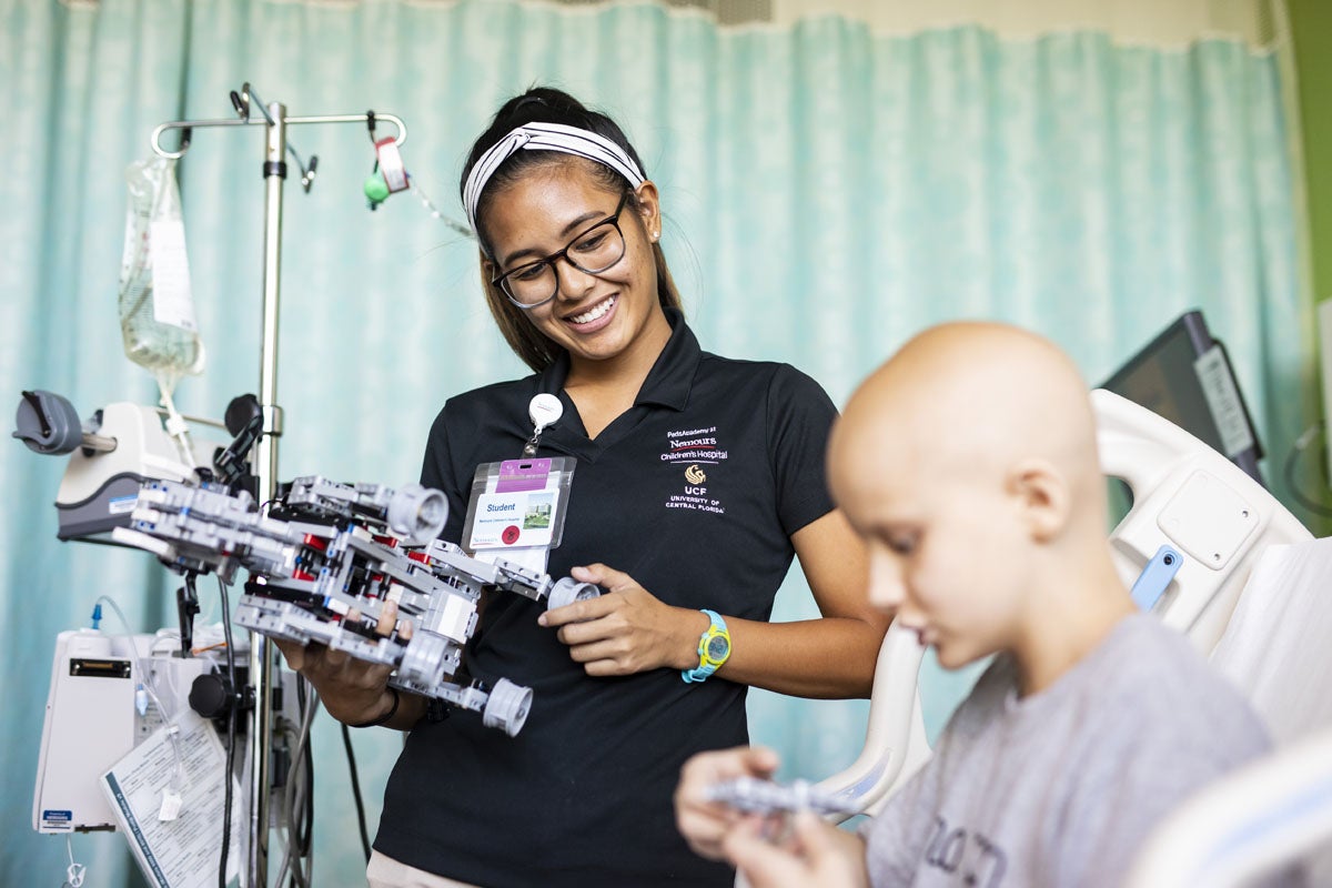 A UCF student volunteer smiles and holds a clipboard while watching a hospitalized child play.