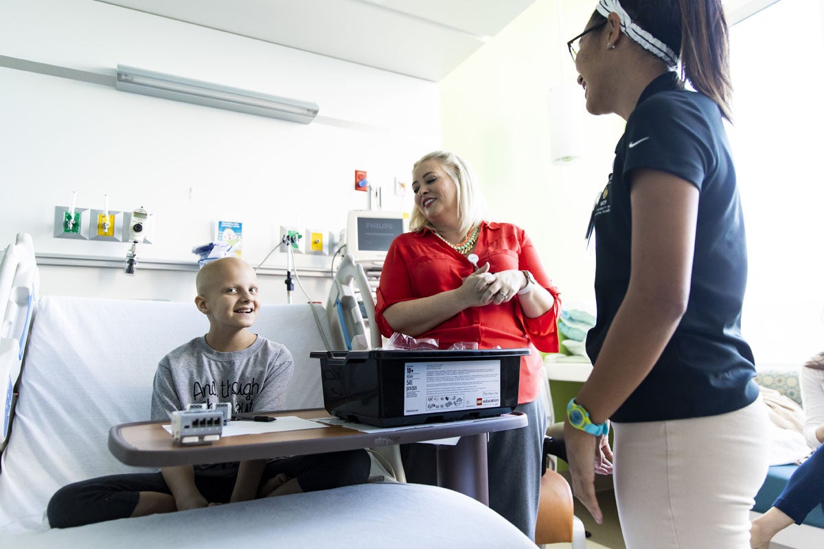 A young child in a hospital bed speaks with a woman and female UCF student.