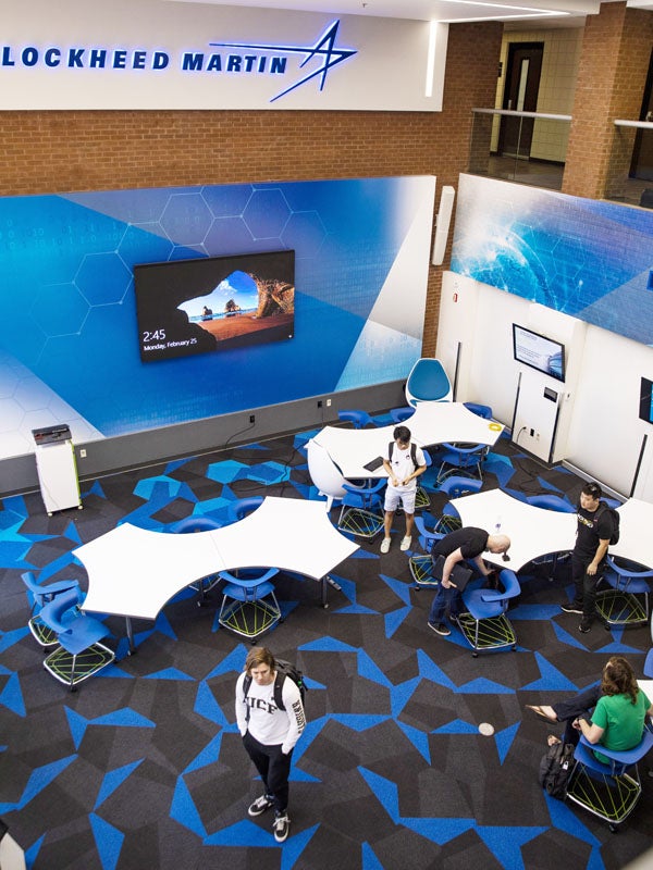 An overhead shot shows a students standing in a room with colorful carpet, whiteboard tables and a TV mounted on the wall.