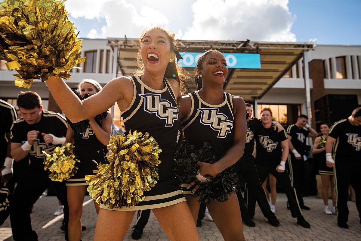 UCF cheerleaders smile while holding black and gold pom-poms outside Millican Hall.