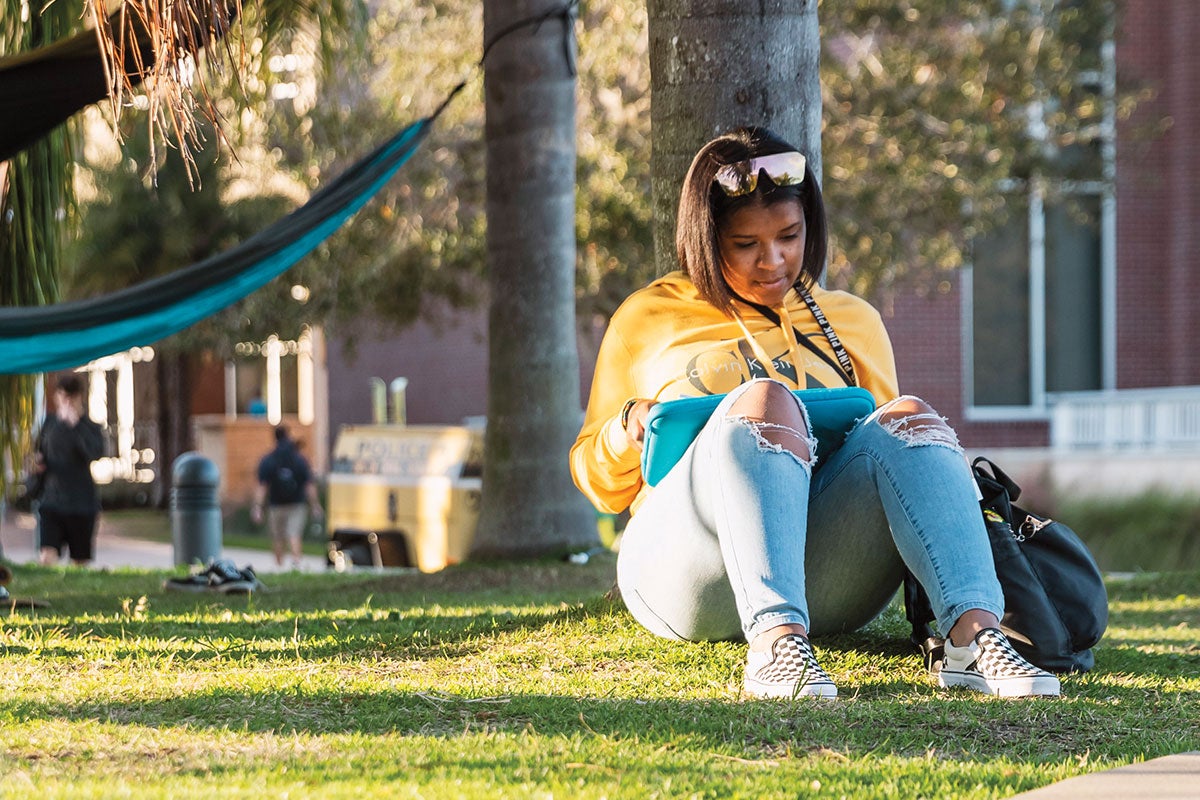 A student studies while sitting under a tree with a hammock hanging in the background.