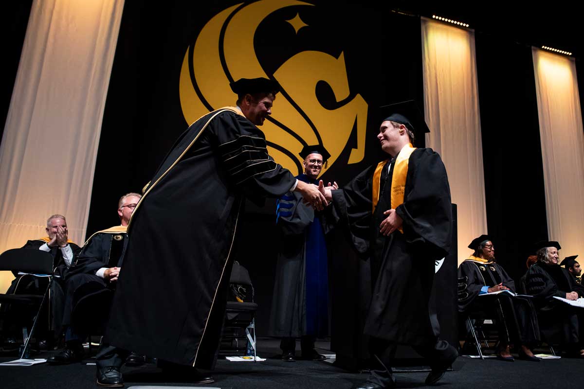 UCF student in cap and gown shakes the hand of a man on stage