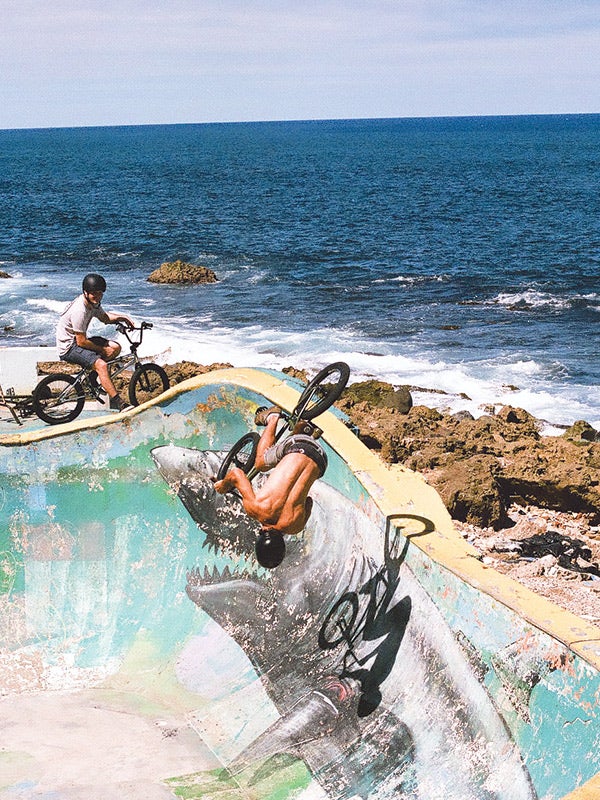 Two men ride BMX bikes in a concrete bowl by the ocean.