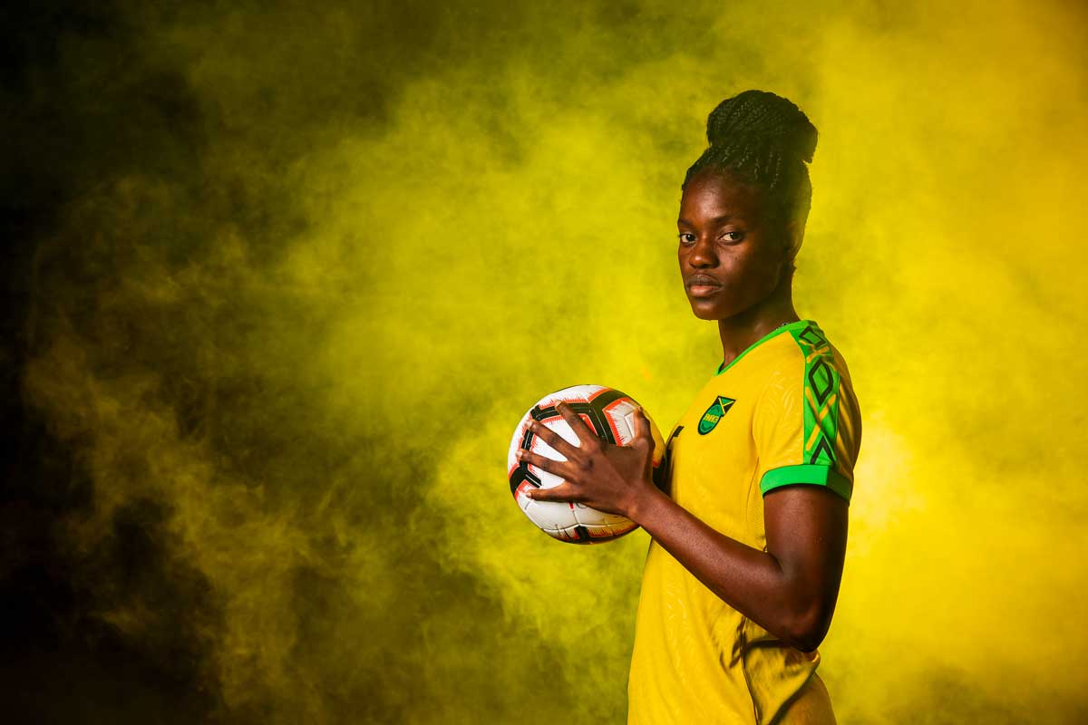 Female soccer player in yellow Jamaica jersey holds soccer ball with yellow smoke behind her
