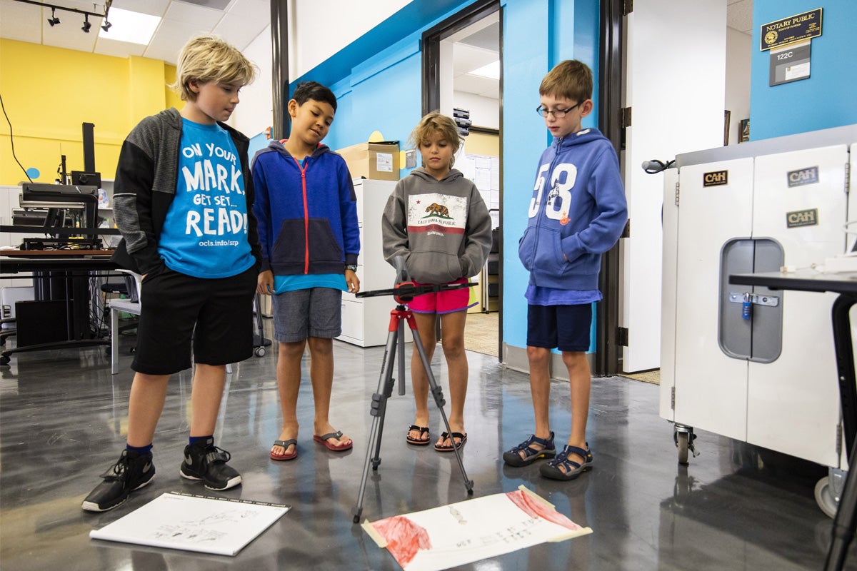 Four kids stand around a mounted tablet set up to take photos.