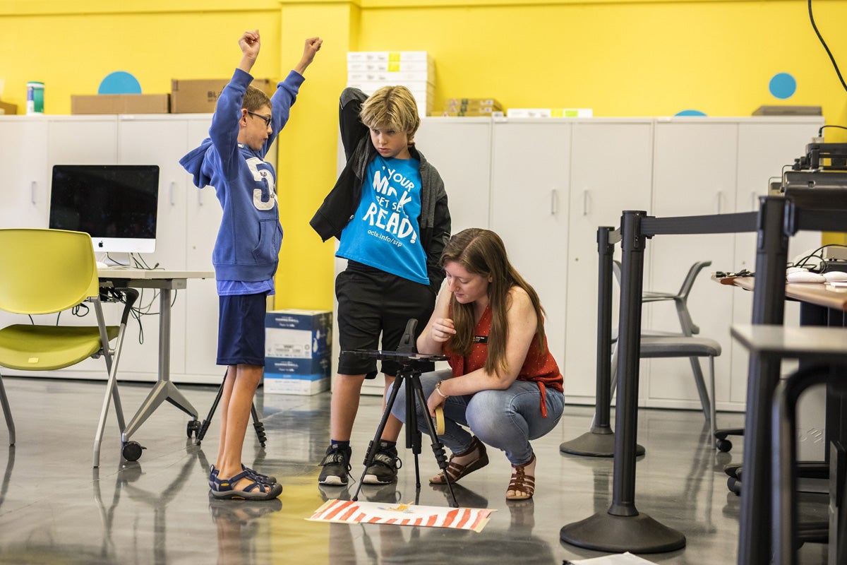 A kid raises his arms while a woman takes a photo of a drawing.