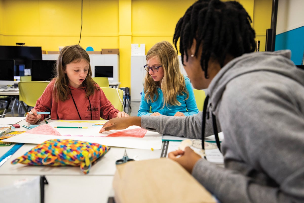 A man speaks to two young girls drawing at a table.