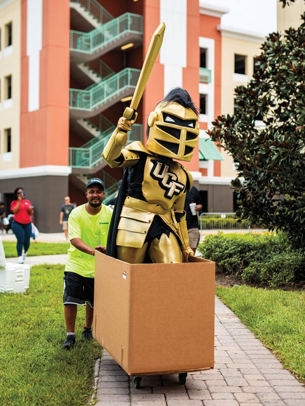 Knightro holds up his sword while standing in a cardboard box being pushed by a mover. UCF Fall 2019