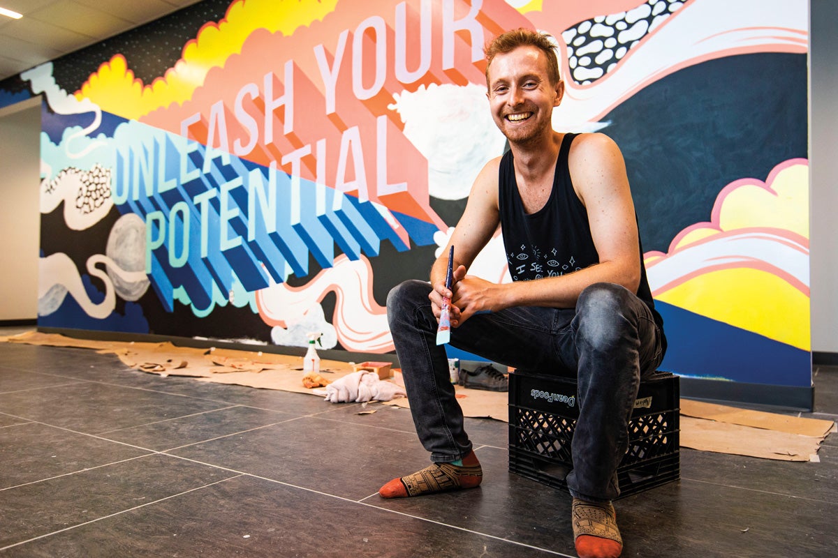 Harry Foreman holds a paint brush in front of a mural that reads 