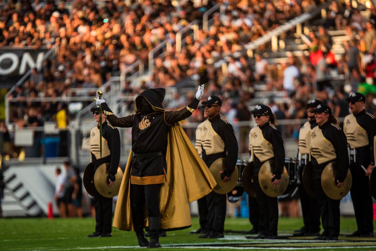 A hooded knight holds a sword while band members holding cymbals stand behind them. 