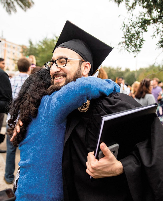 Man wearing graduation cap and gown hugs a woman