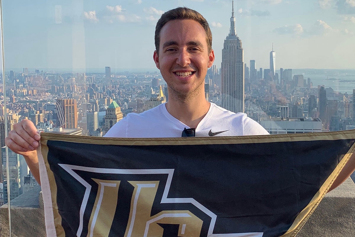 Brandon Aliberti holds a UCF flag while posing for a photo with a skyline behind him.