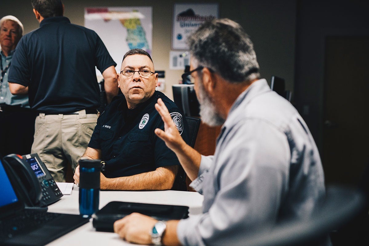 A male police officer and another man speak while sitting.