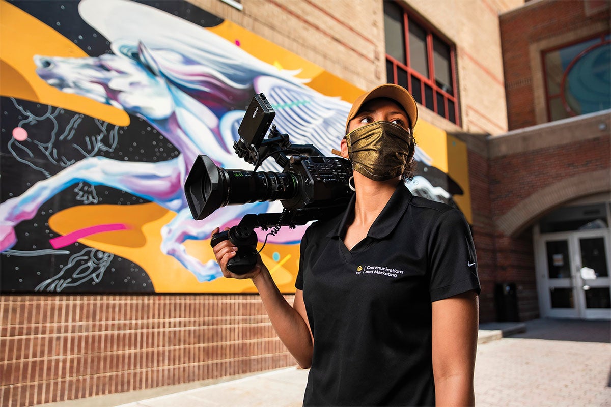 Jasmine Kettenacker wears a mask while holding a camera in front of the Pegasus mural.