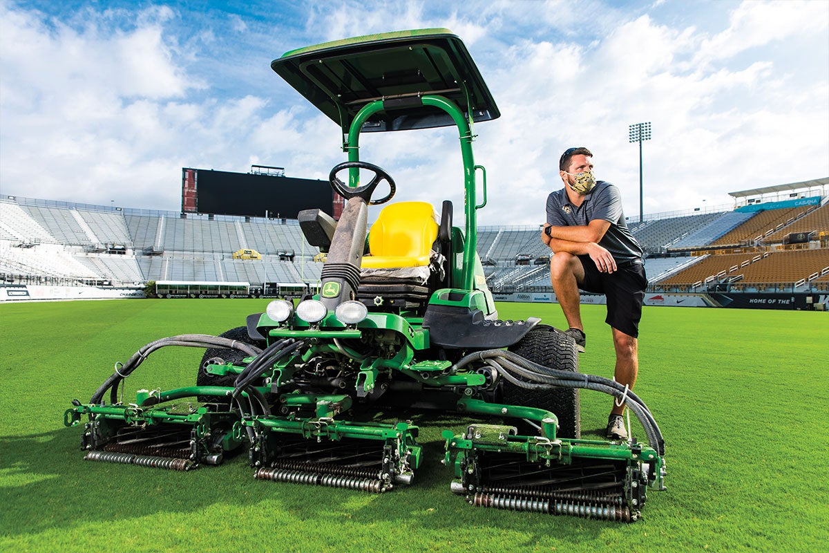 Jon Palmer wears a mask and poses on a piece of landscaping gear while instead Spectrum Stadium.