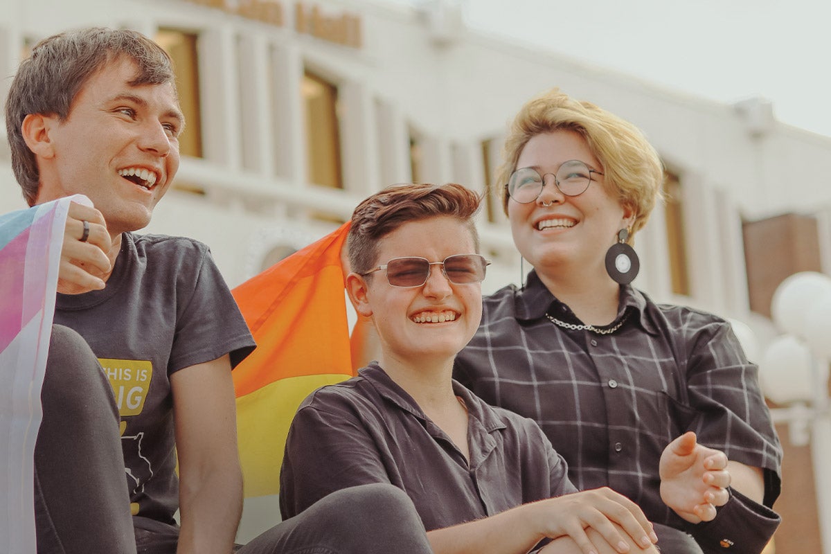 Three students laugh while holding various pride flags