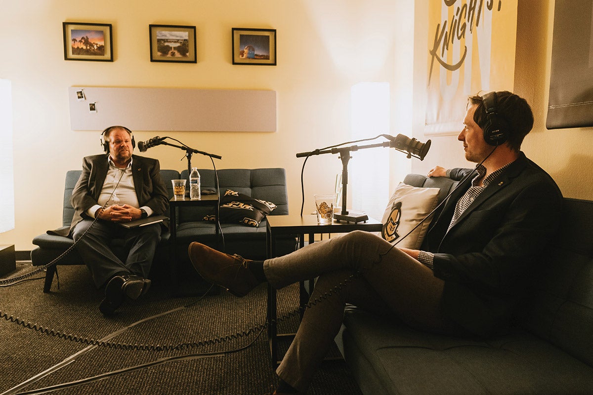 Alexander N. Cartwright and Alex Cumming sit on coaches while wearing headphones in front of mics.