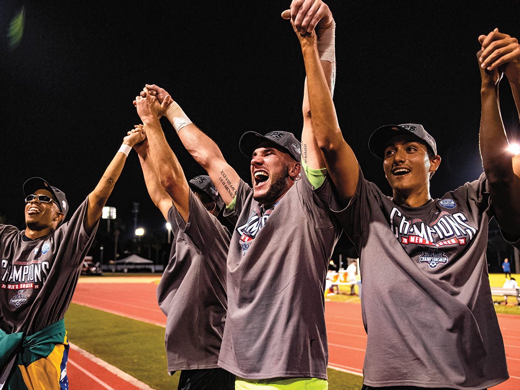 Four members of the men's soccer team clasp hands and raise arms above their heads in celebration