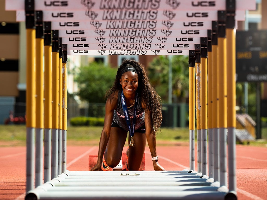 Portrait of Rayniah Jones in the starting blocks, framed by a tunnel of hurdles lined up in front of her