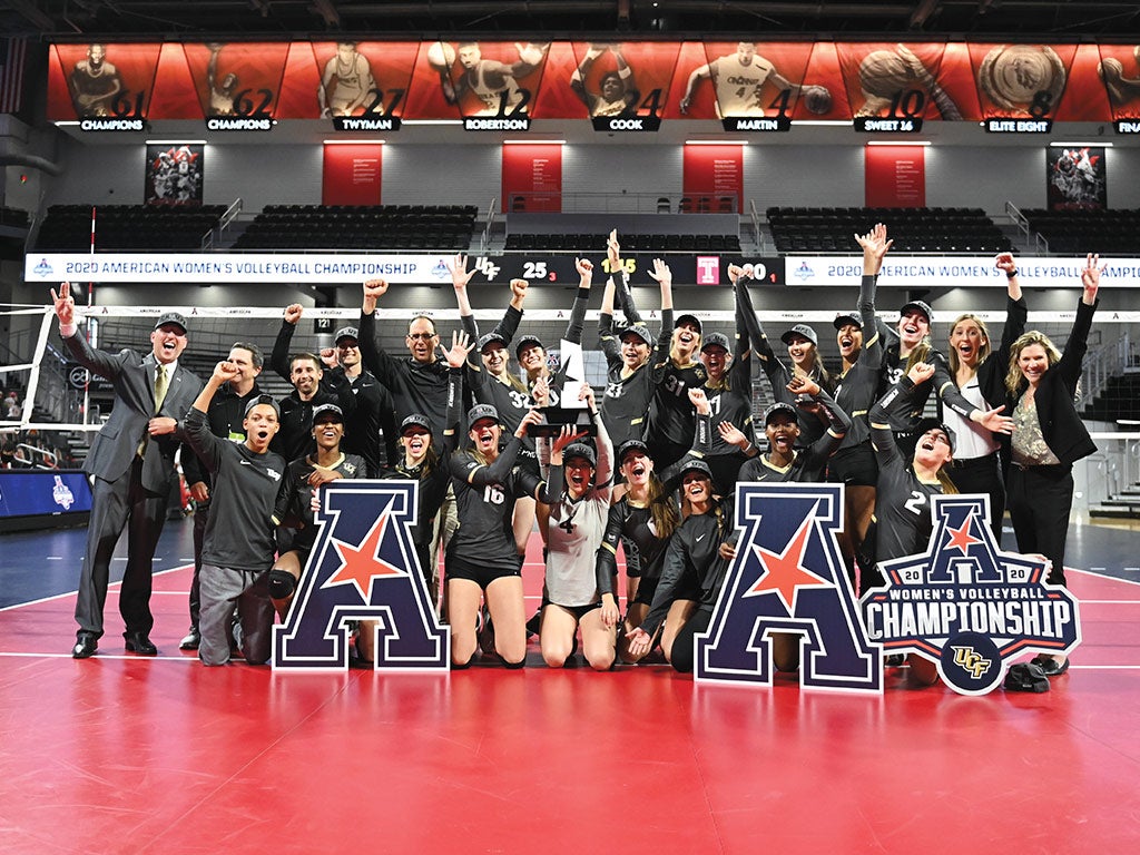 Volleyball team gathers for a photo in front of the net on the court while holding American Athletic Conference champions signs