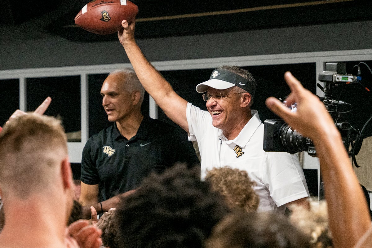 Guz Malzahn holds up a football in the locker room while Terry Mohajir stands nearby.