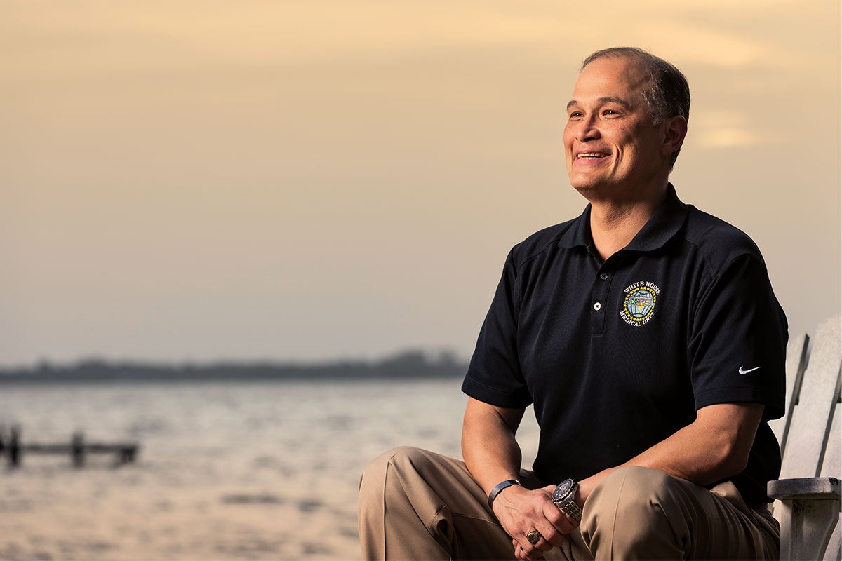 Portrait of Tom Weichart, wearing a black Nike polo shirt and khaki pants, sitting near lake at sunset